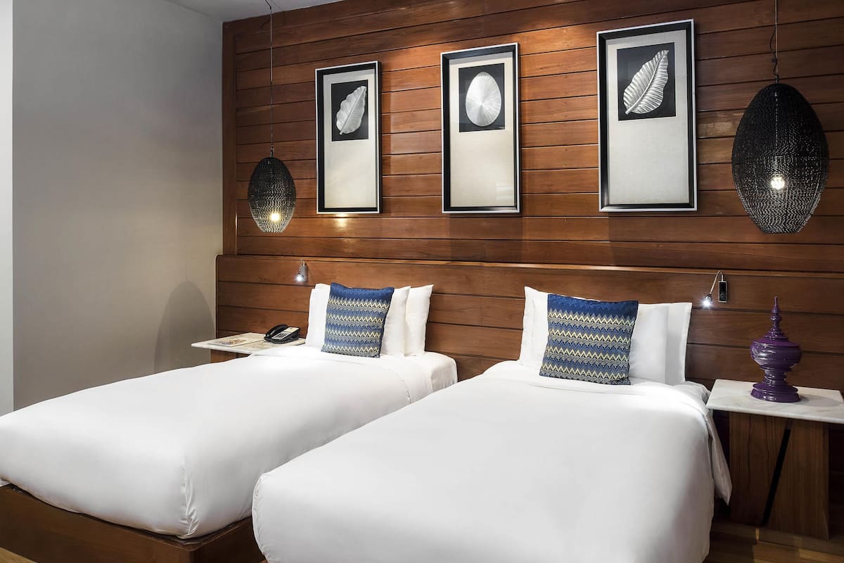 A hotel room with two beds, wooden headboard, framed artwork, bedside lamps, and a nightstand with a lamp.
