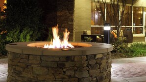A stone fire pit with flames, surrounded by a stone wall and greenery, in front of a building with large windows and a seating area.