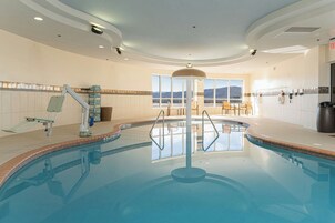 Una piscina interior con trampolín, un espacio soleado con mesa y sillas, y vistas a las montañas a través de grandes ventanales.