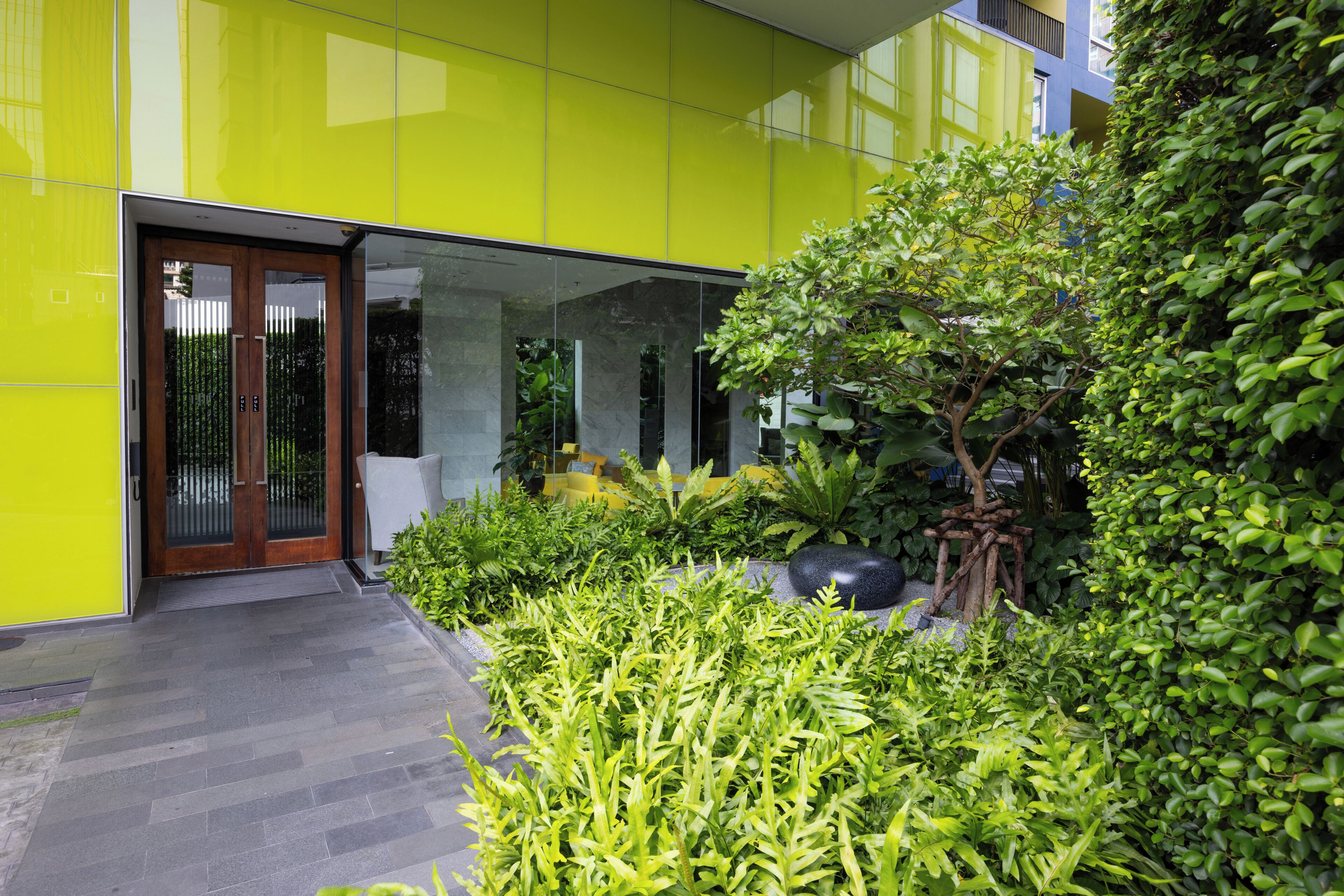 Un edificio moderno con una facciata giallo brillante, un ingresso in vetro e un giardino curato con diverse piante e un albero.