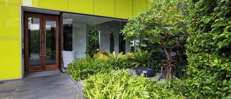Un edificio moderno con una facciata giallo brillante, un ingresso in vetro e un giardino curato con diverse piante e un albero.
