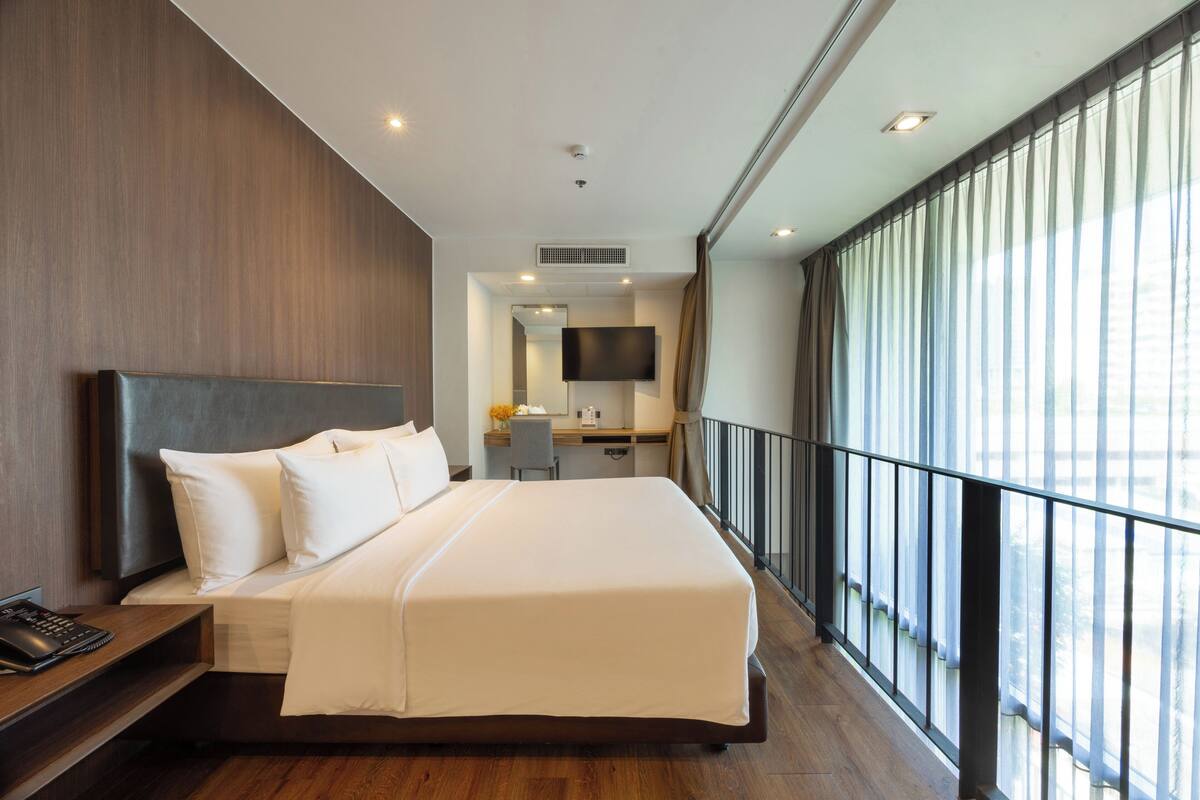 A modern hotel room with a large bed, a desk, a chair, a flat-screen TV, and a balcony with a railing.