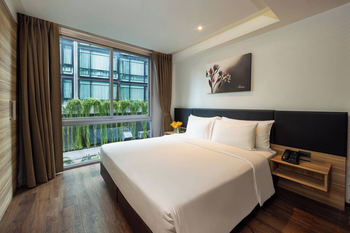 A hotel room with a large bed, a bedside table with a phone, a window with curtains, and a painting on the wall.