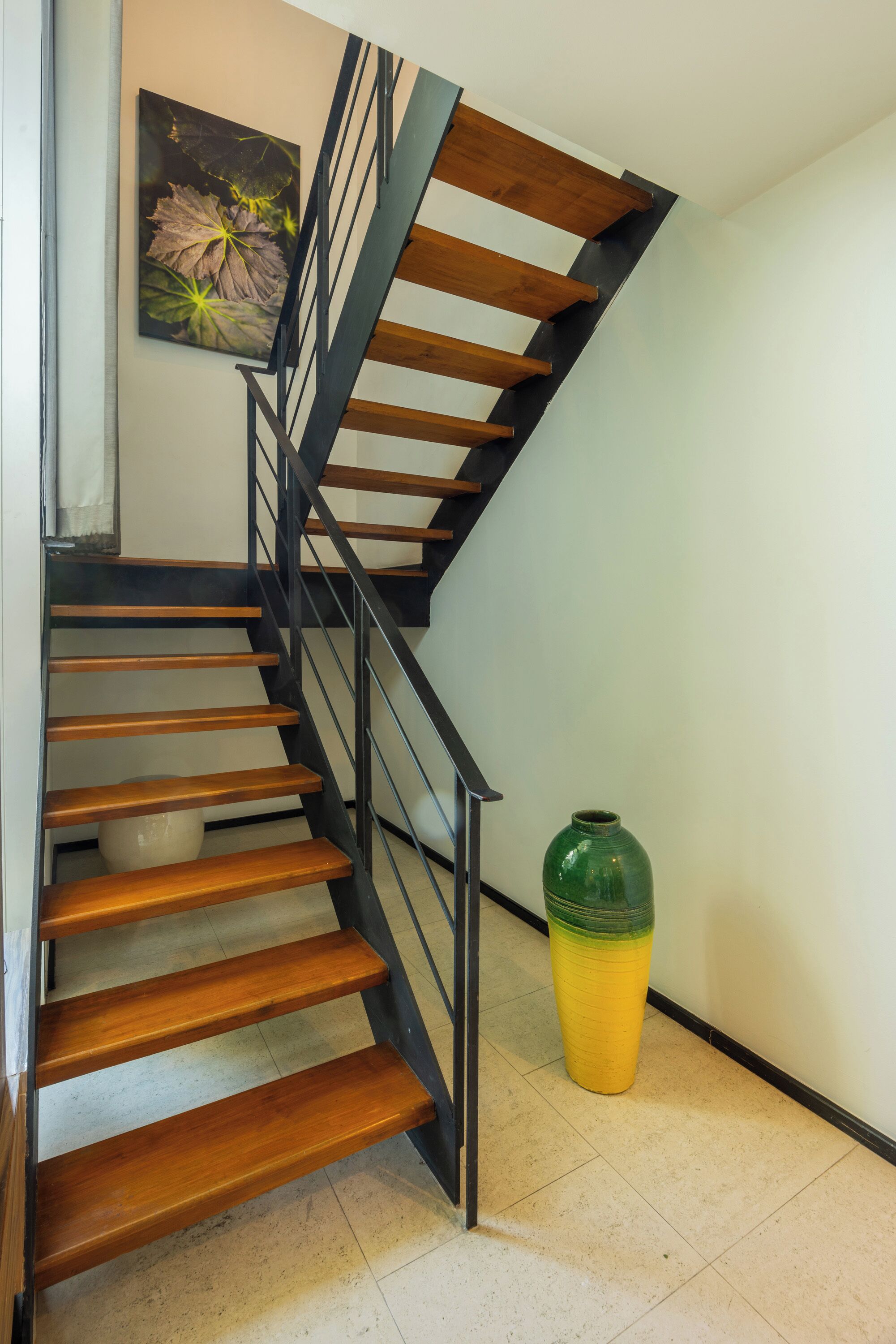 Un escalier avec des marches en bois et des rampes noires, un vase posé sur le sol et un tableau accroché au mur.
