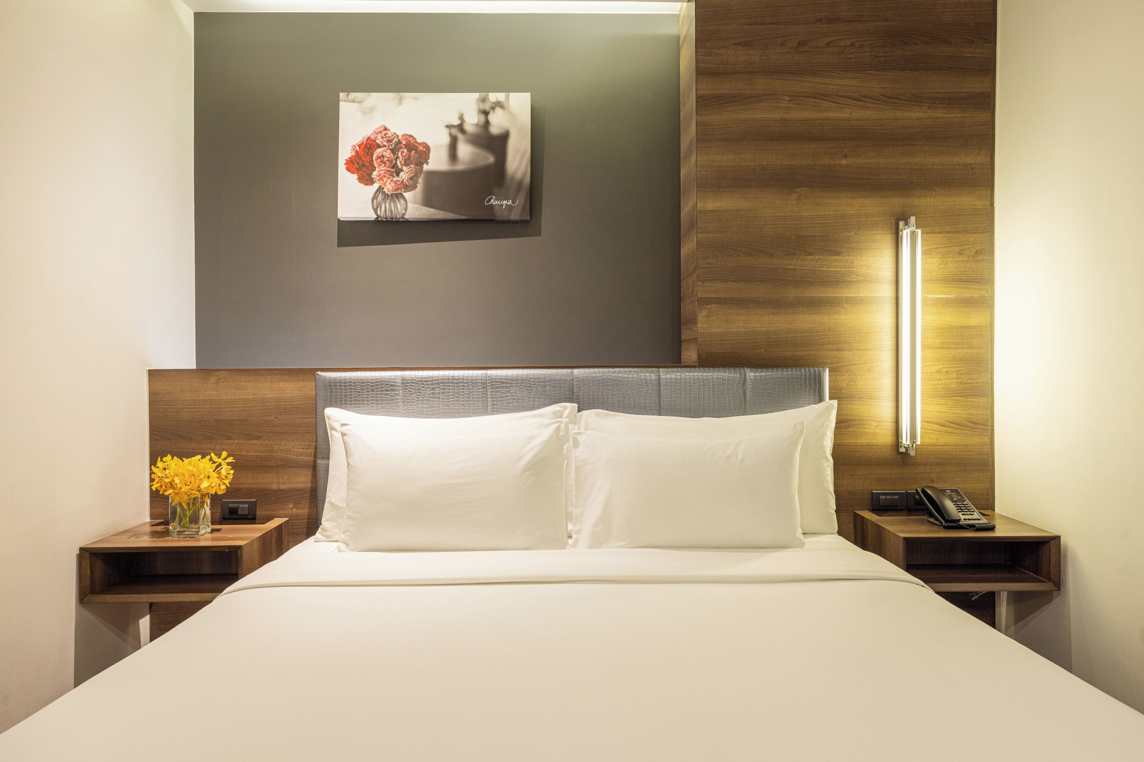 A hotel room with a bed, two bedside tables, a nightstand with a vase of flowers, a wall-mounted picture, and a bedside lamp.