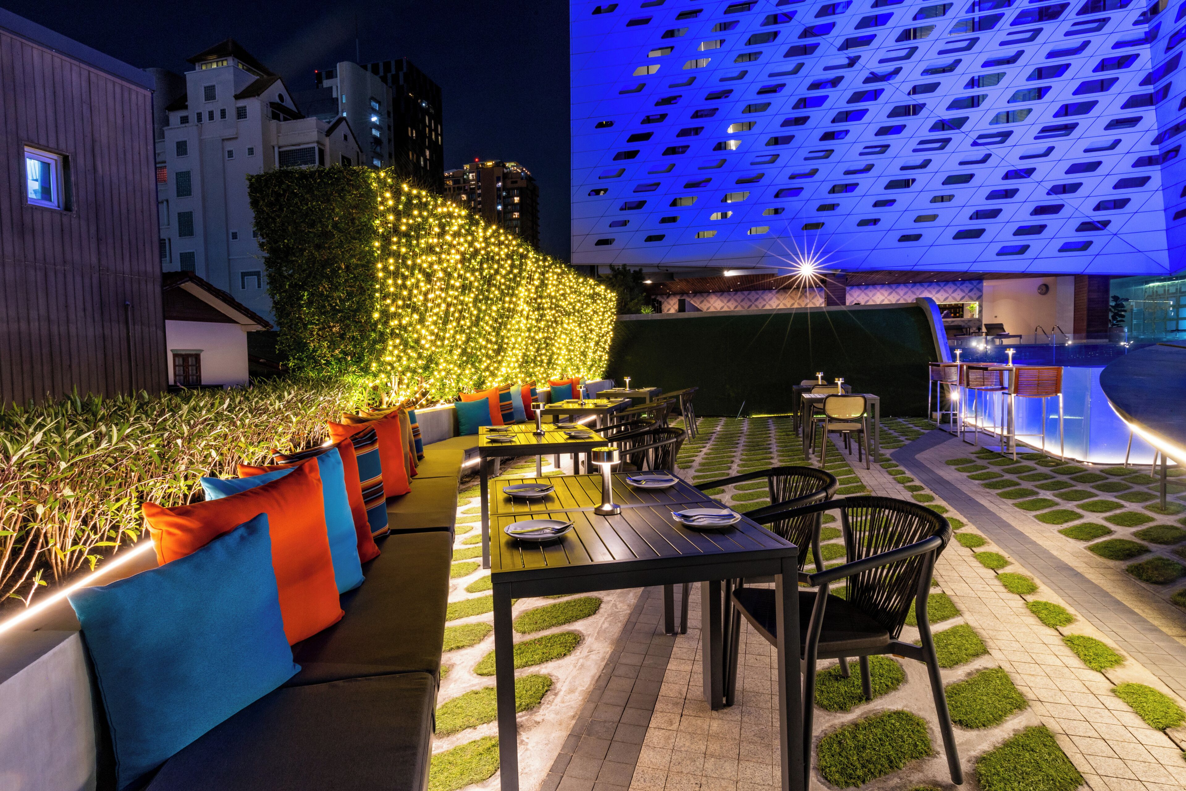 An outdoor dining area with tables and chairs, surrounded by greenery and illuminated by ambient lighting.