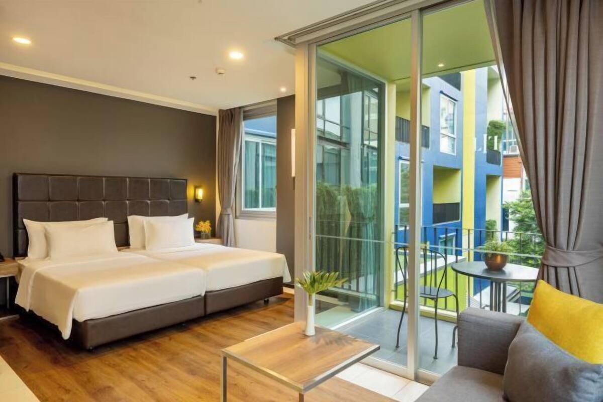 A modern hotel room with a large bed, a sofa, a small table, and a balcony with a view of the city.