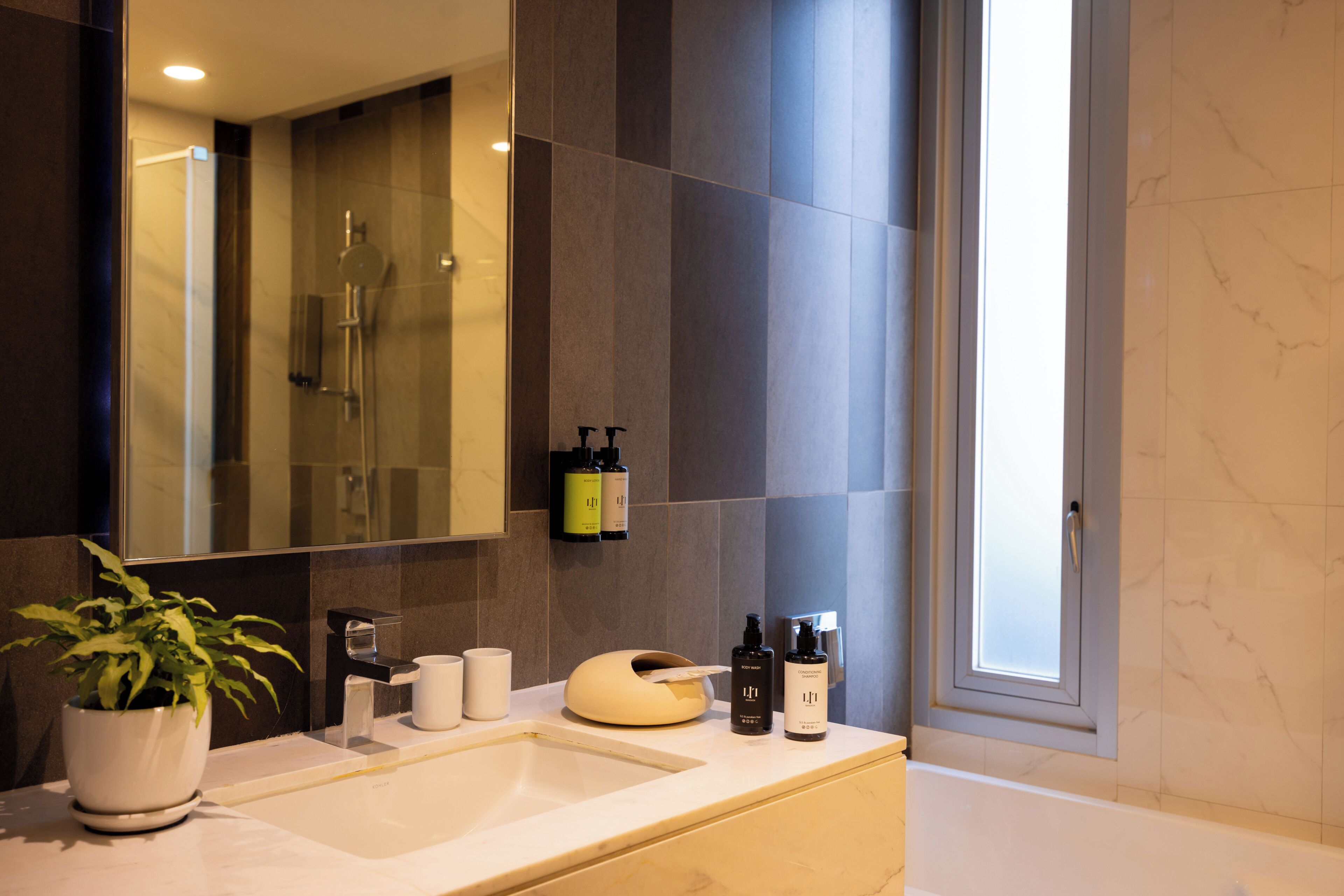 A modern bathroom with a large mirror, a sink, a potted plant, and toiletries on the counter.