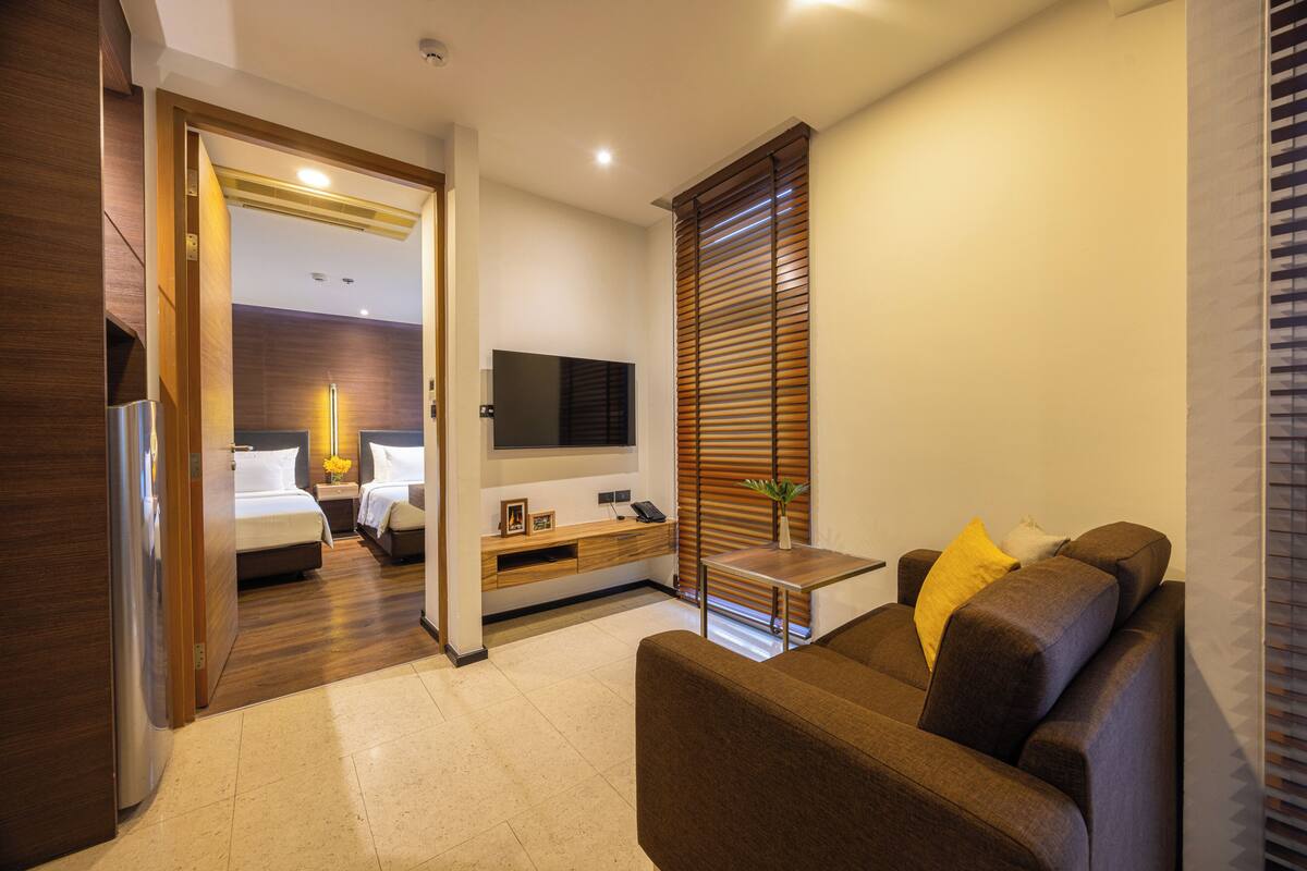 A modern hotel room with a sofa, a TV, a bedside table, a wooden floor, and a door leading to a bathroom.
