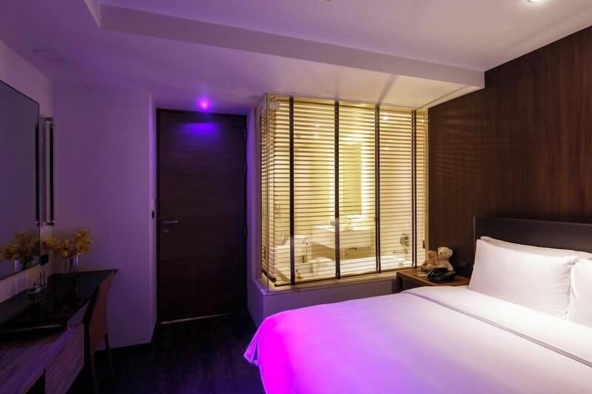 Una habitación de hotel moderna con cama, mesita de noche y un gran ventanal con persianas.