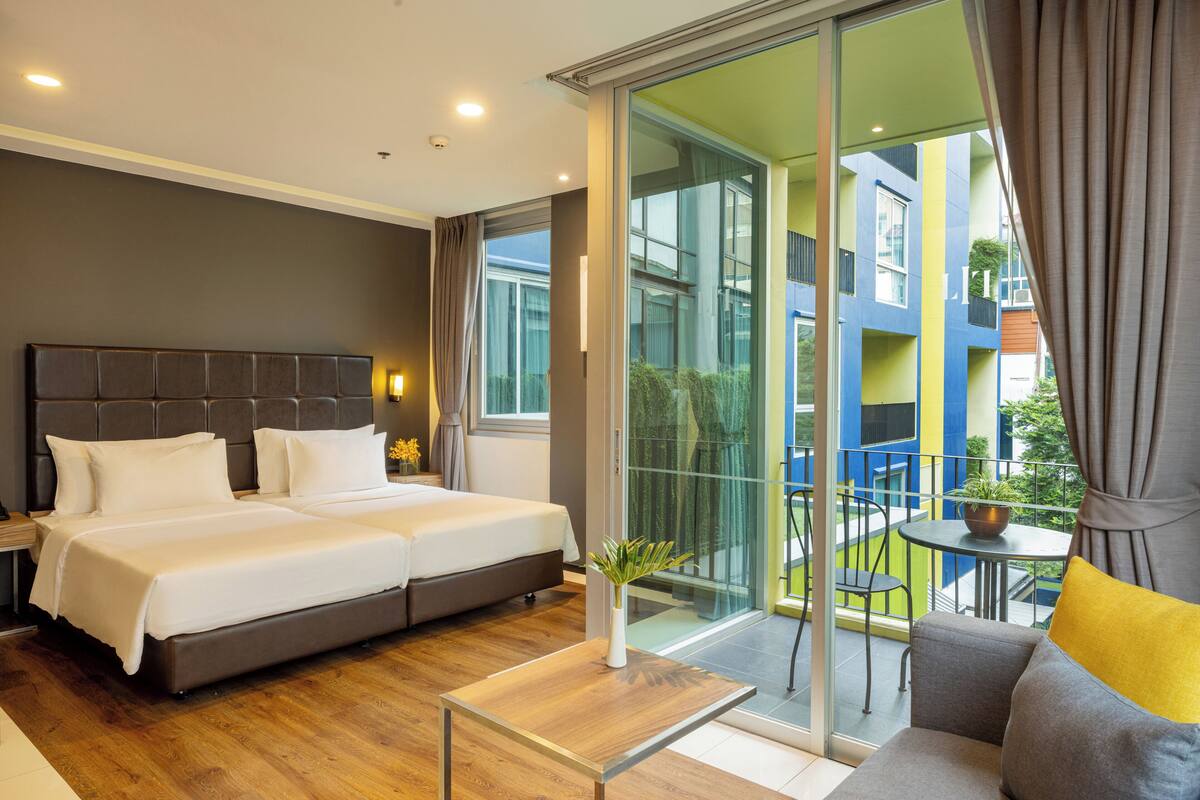 A modern hotel room with a large bed, a small table, a sofa, and a balcony with a view of the city.
