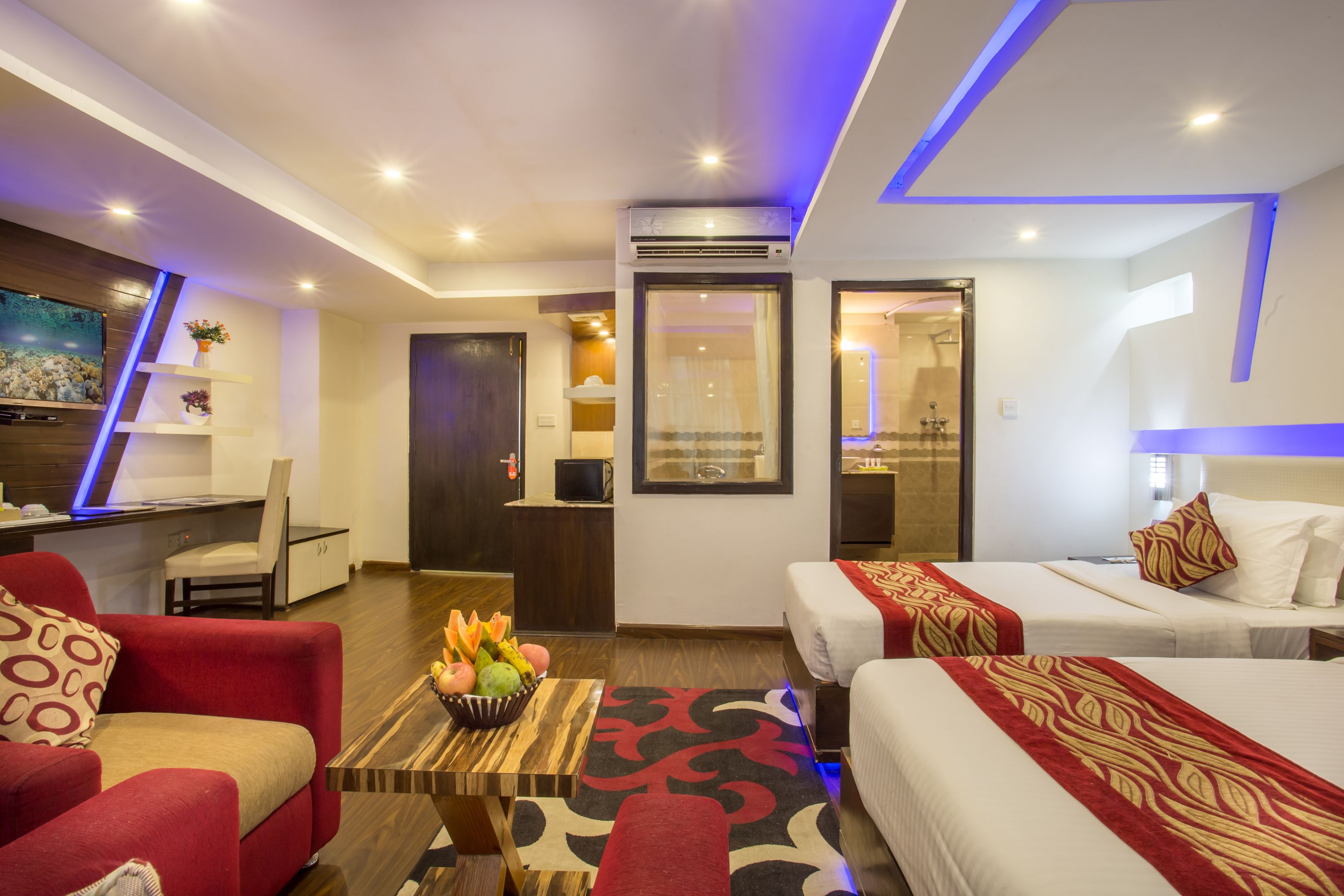 A hotel room with two beds, a desk, a TV, and a seating area with a table.