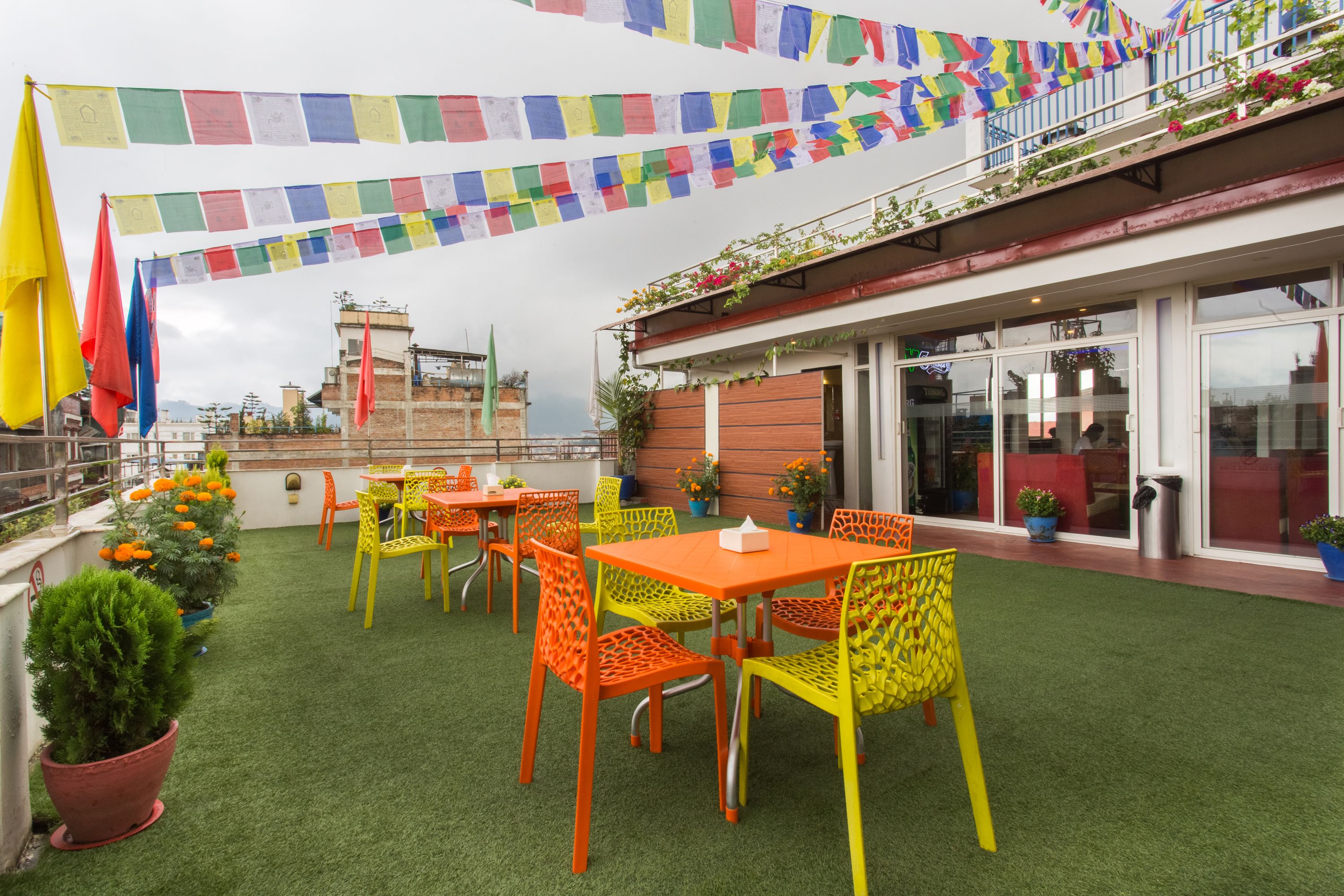 A rooftop terrace with green artificial turf, orange and yellow chairs, and colorful prayer flags.