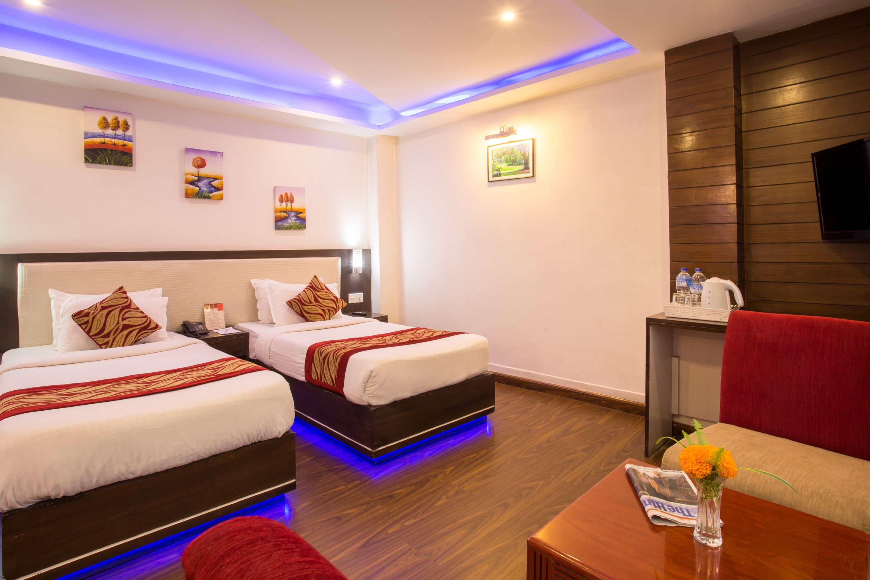 A hotel room with two beds, a TV, a small table with a vase, and artwork on the walls.