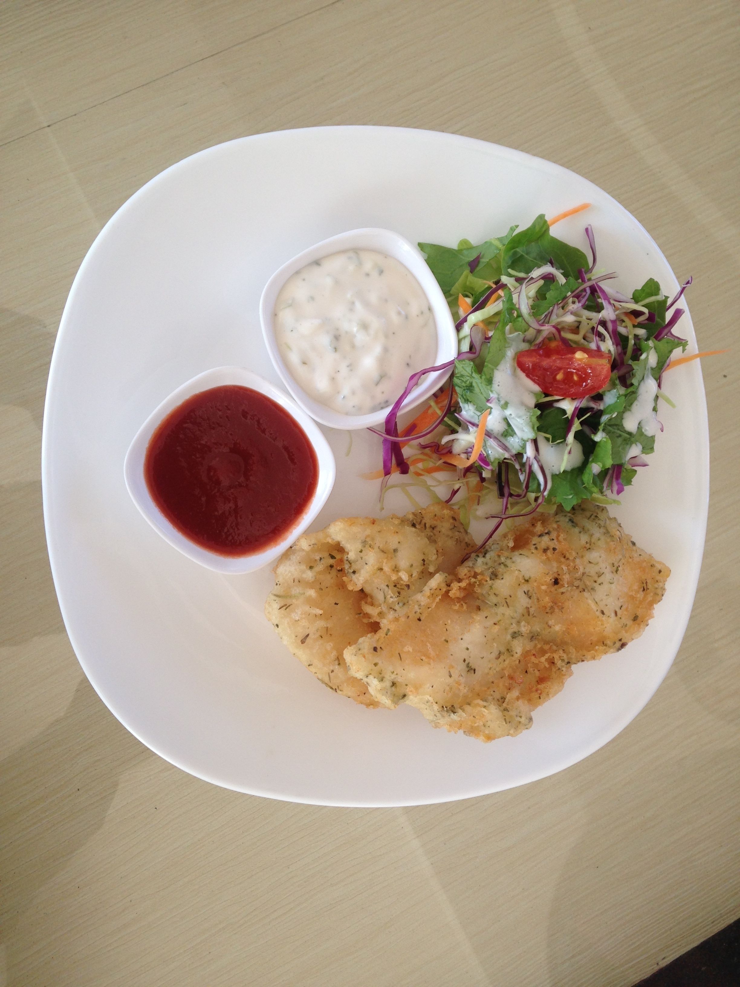 A white plate with a piece of breaded fish, a salad, and two small bowls of sauce.