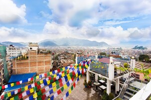 Un paisaje urbano con vistas a montañas distantes, cielo azul y coloridas banderas de oración.