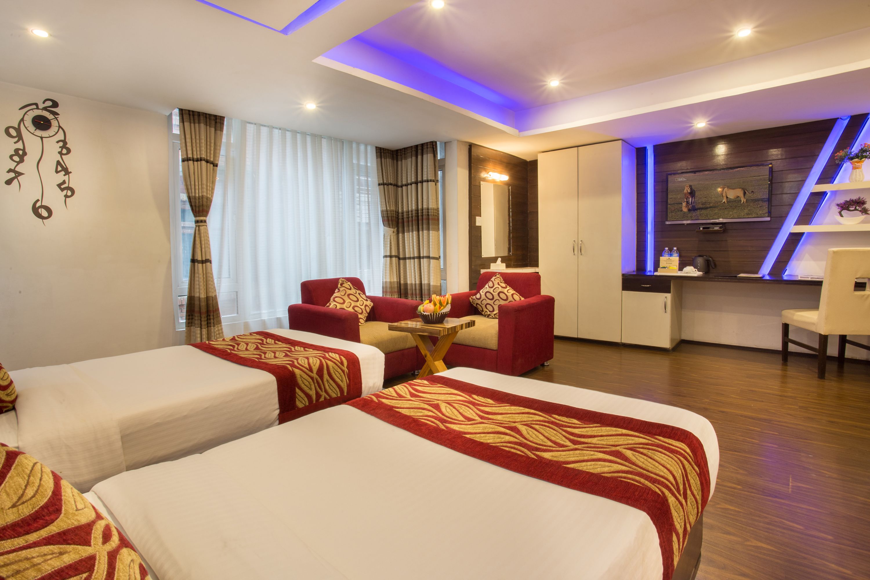 A hotel room with two beds, a small table with a bowl of fruit, a red sofa, a TV, and a minibar.