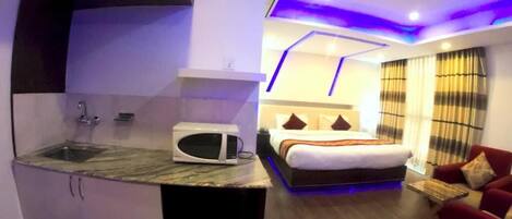A compact hotel room with a kitchenette, a bed with a headboard, a red sofa, and a window with curtains.