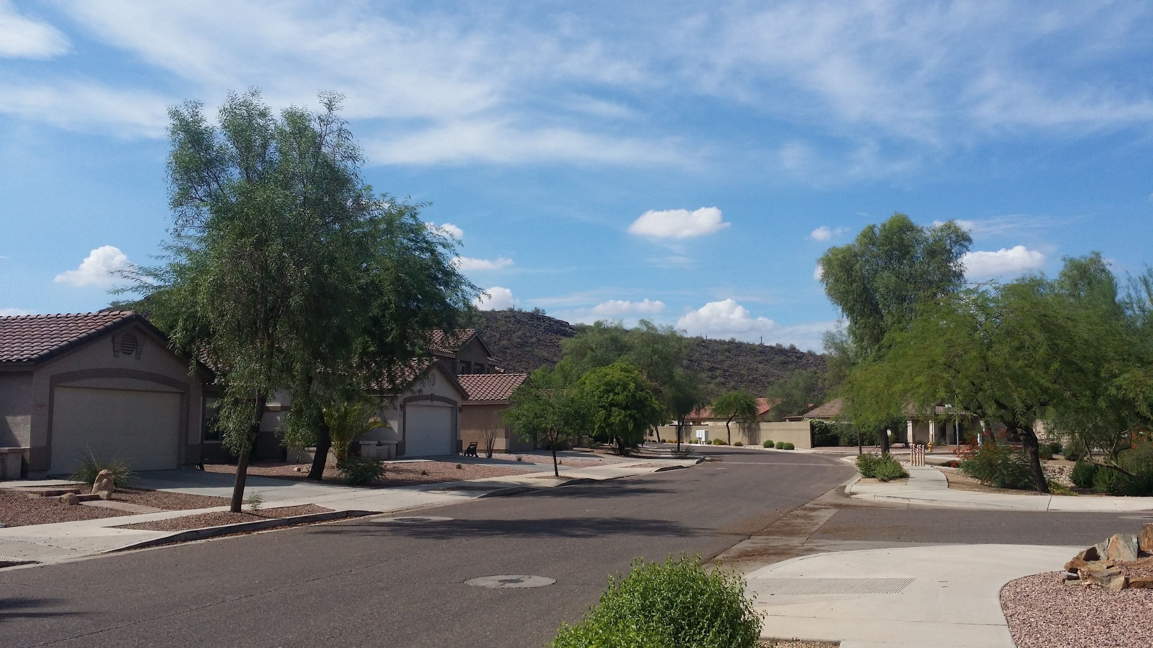 A quiet suburban street lined with single-family homes, featuring a clear sky and a hilly backdrop.
