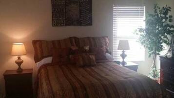 A bedroom with a bed, two bedside lamps, a window with blinds, and a potted plant.