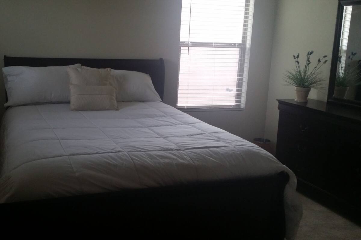 A neatly made bed with white bedding and a dark headboard in a room with a window and a potted plant.