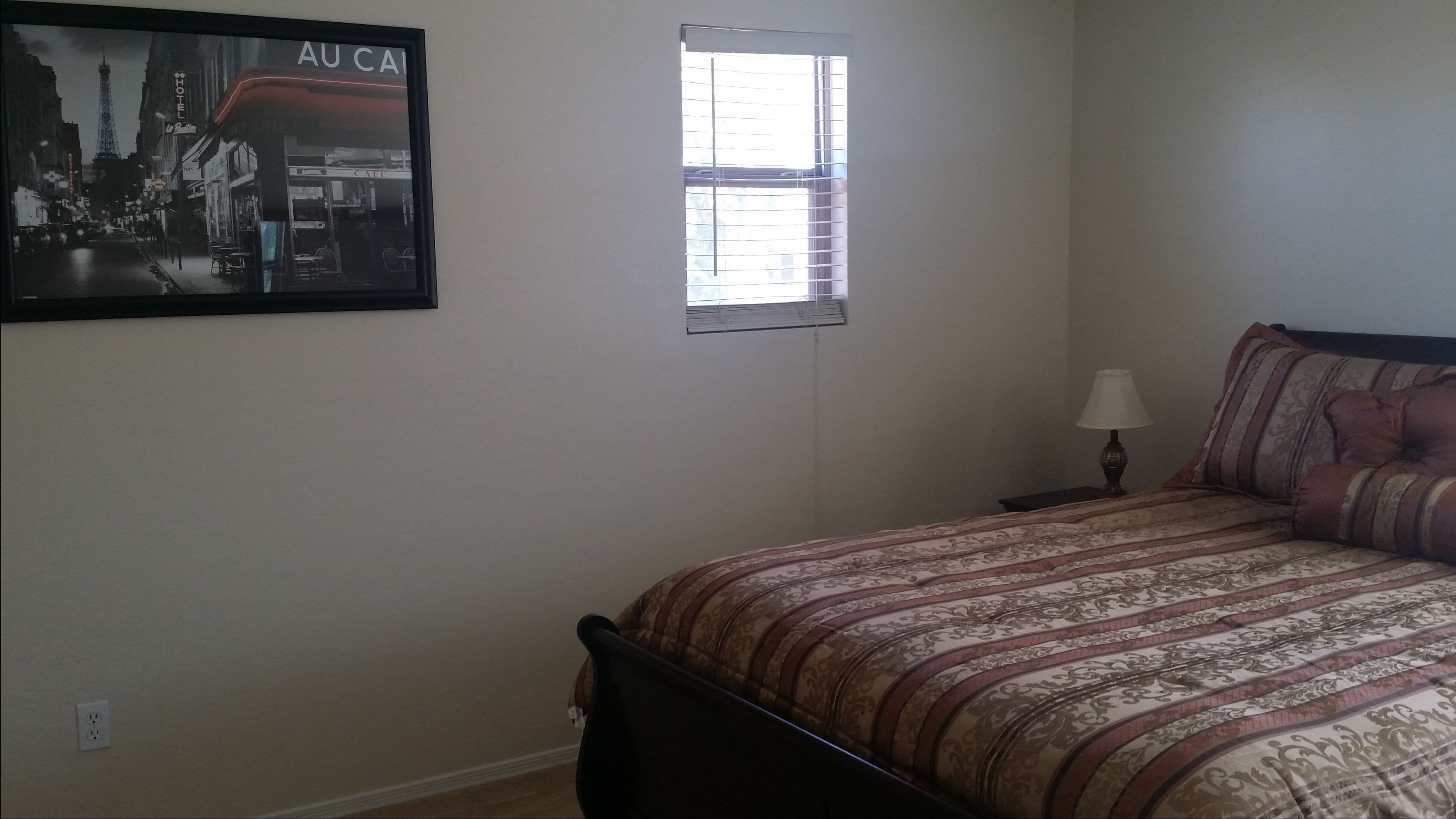 Une chambre à coucher comprenant un lit, une table de chevet, une lampe, une fenêtre et un tableau représentant un paysage urbain accroché au mur.
