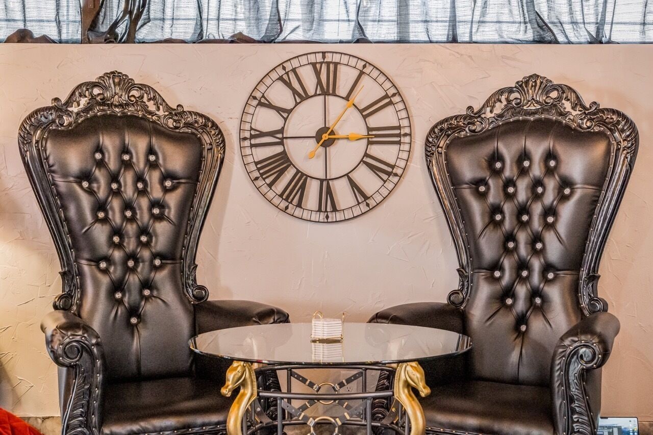 Two ornate leather armchairs with a tufted design, a round table with a glass top, and a large wall clock.