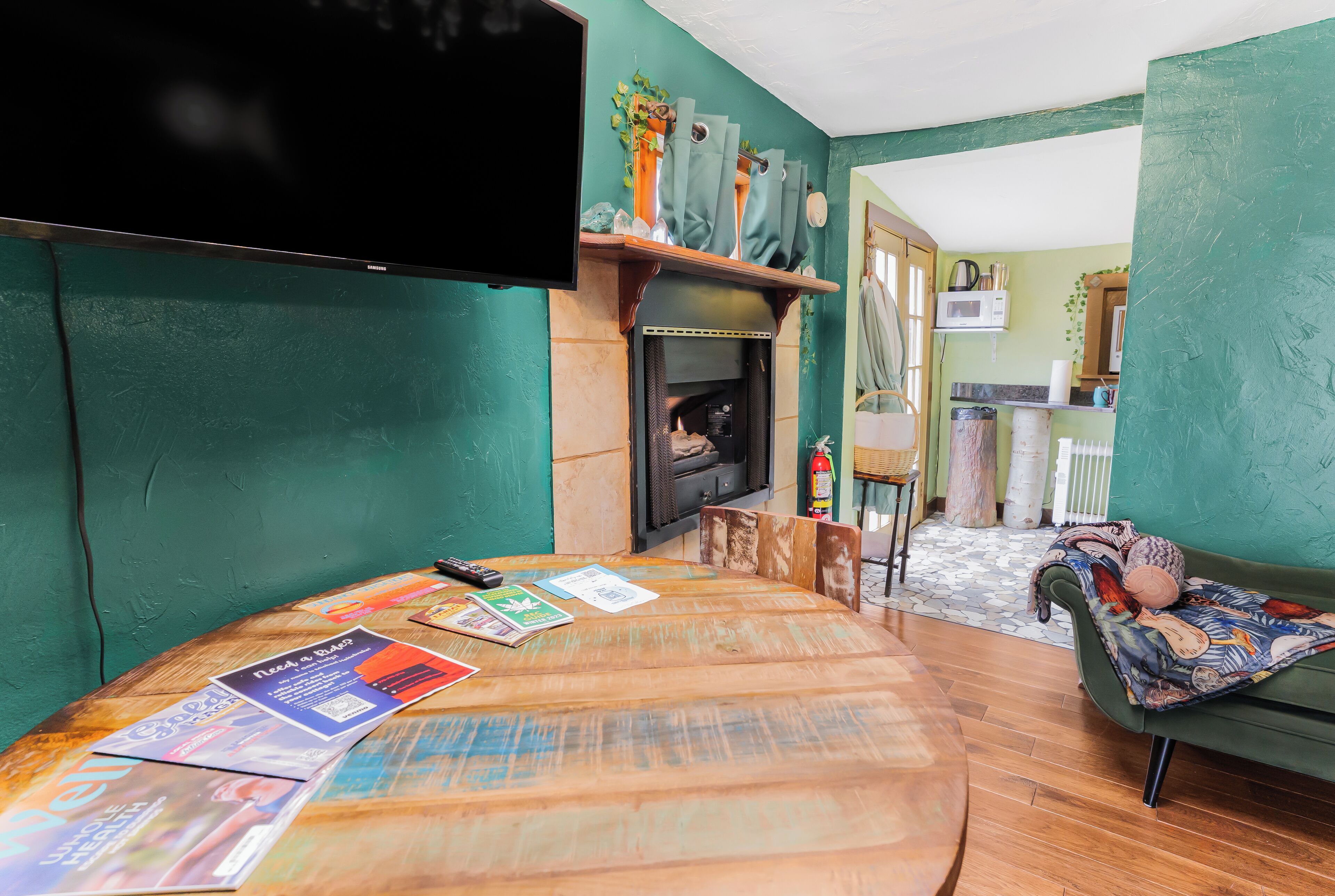 A cozy living room with a fireplace, a round wooden table with magazines, a green sofa, and a television mounted on the wall.