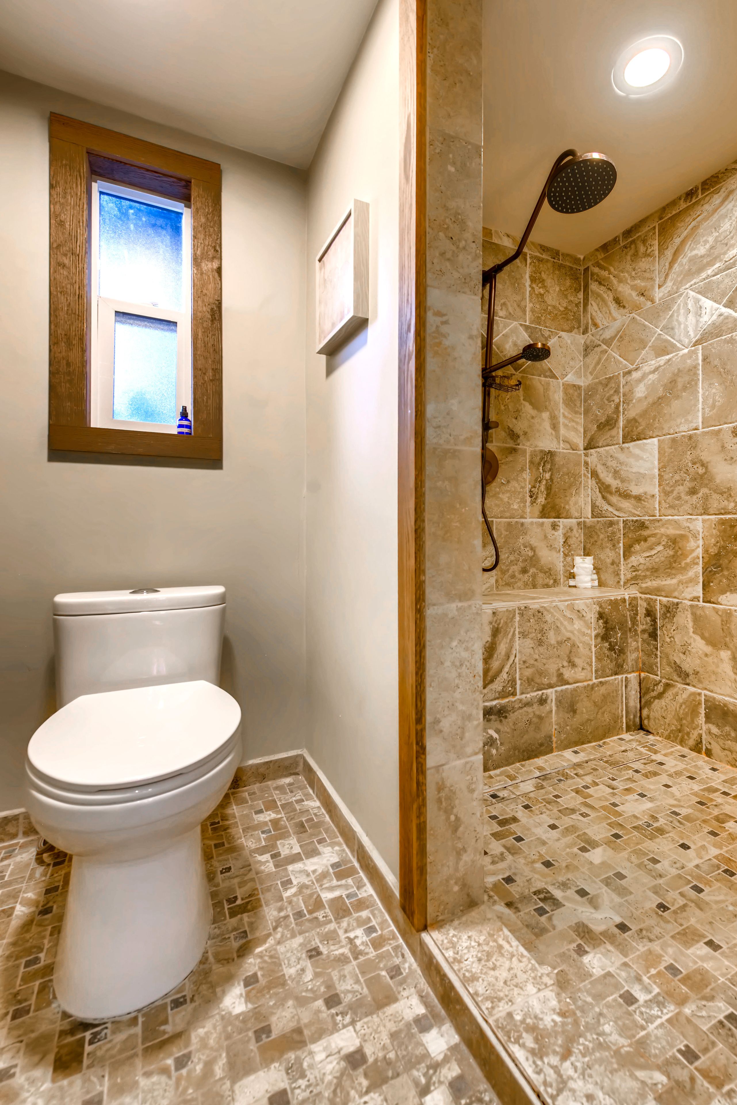 A bathroom with a toilet, a shower with a handheld showerhead, and a window above the toilet.
