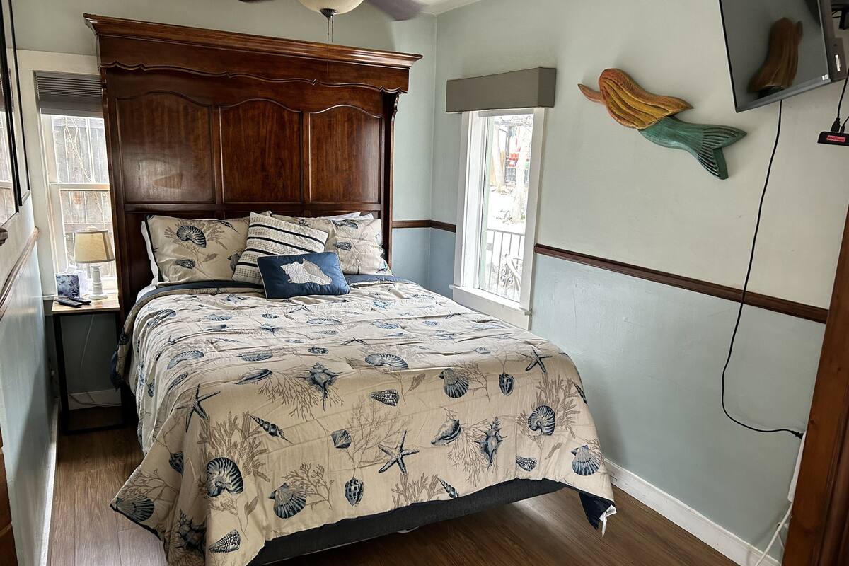 Een slaapkamer met een houten hoofdbord, een bed met een bloemenprint sprei, een nachtkastje met een lamp en een raam met uitzicht.