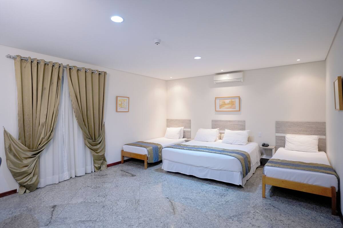 Une chambre d’hôtel avec deux lits simples, une grande fenêtre avec des rideaux beiges et un sol en marbre.