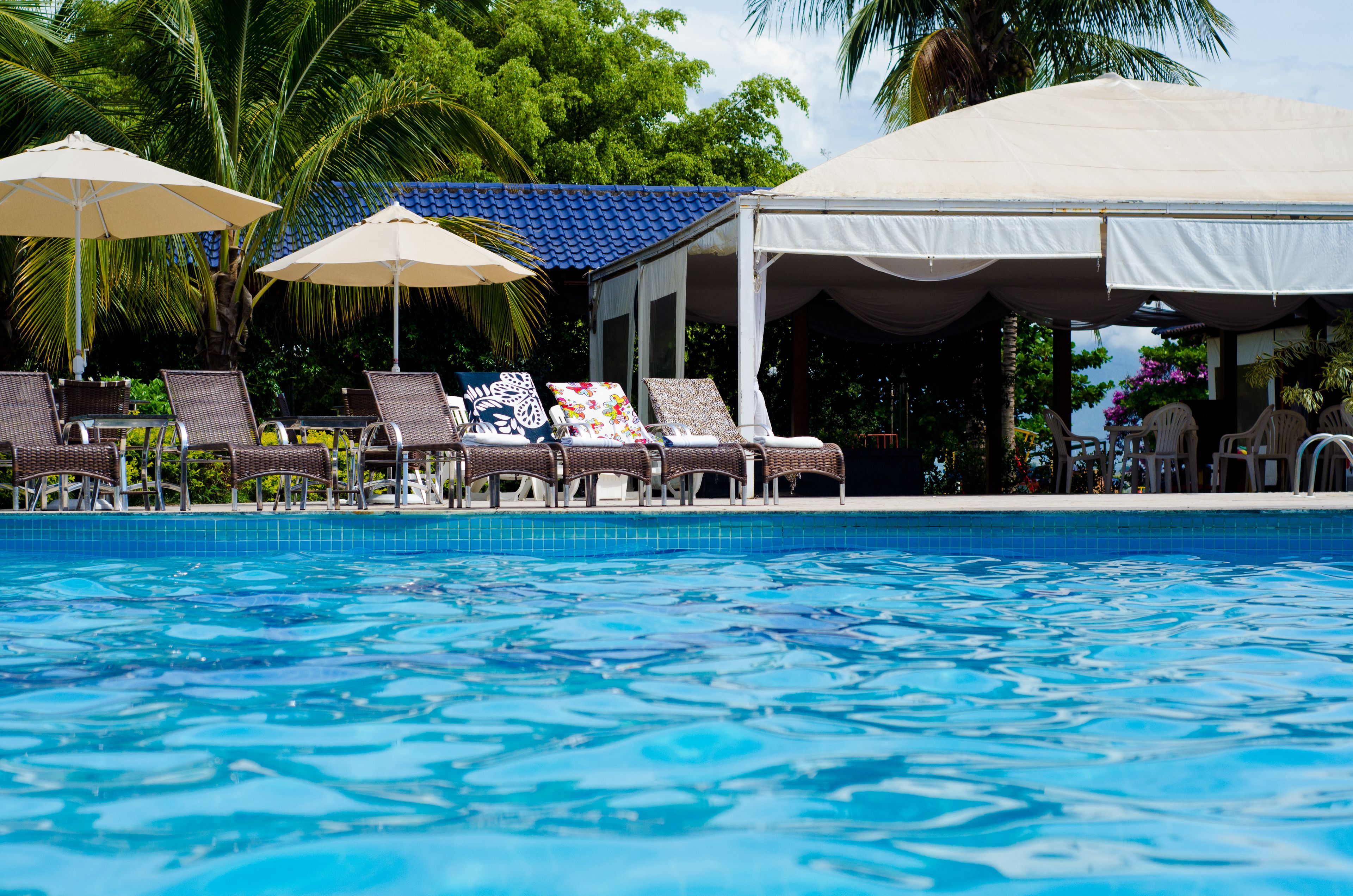 A pool area with lounge chairs and umbrellas, surrounded by palm trees and a covered pavilion.