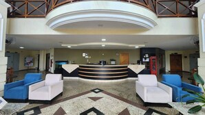 Un vestíbulo de hotel con una mesa de recepción, dos sillones azules, un sillón blanco, un teléfono rojo y un diseño decorativo en el suelo.