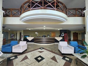 Un hall d’hôtel avec un comptoir d’accueil, deux fauteuils bleus, un fauteuil blanc, un téléphone rouge et un motif décoratif sur le sol.
