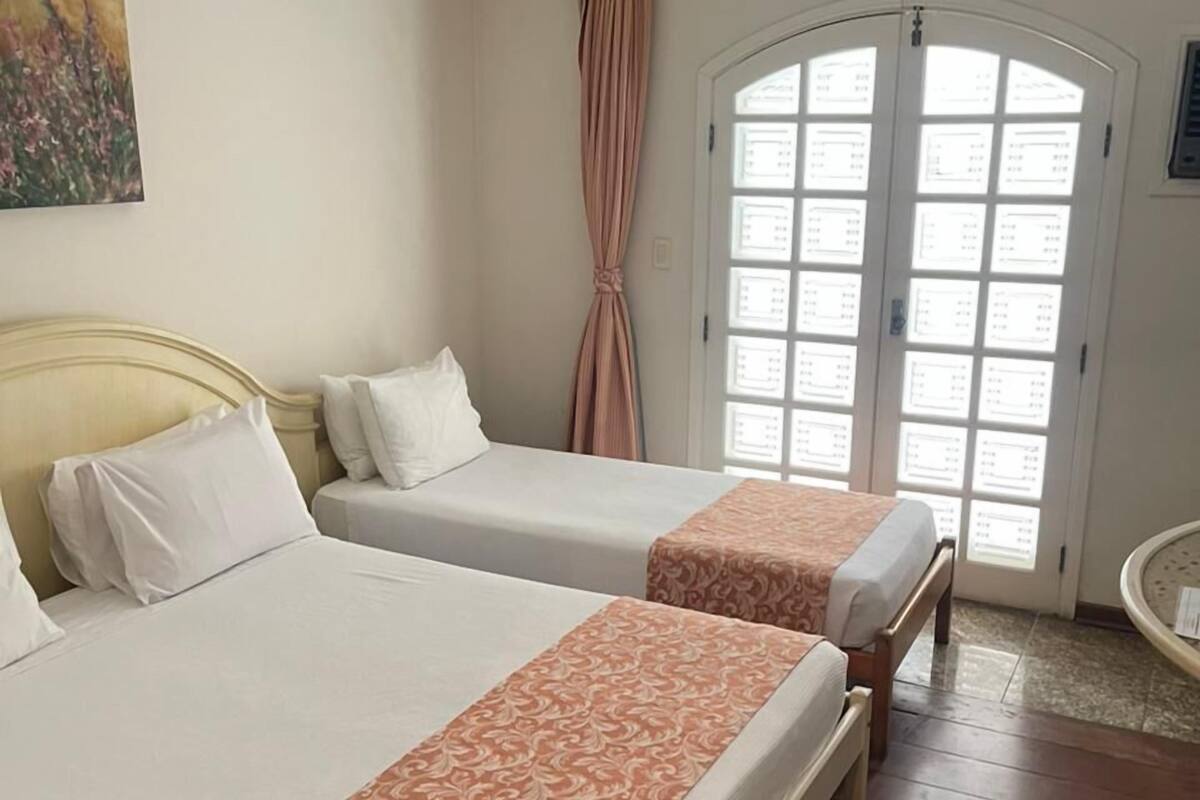Une chambre d’hôtel avec deux lits, une grande fenêtre avec des rideaux, un tableau au mur et un plancher en bois.