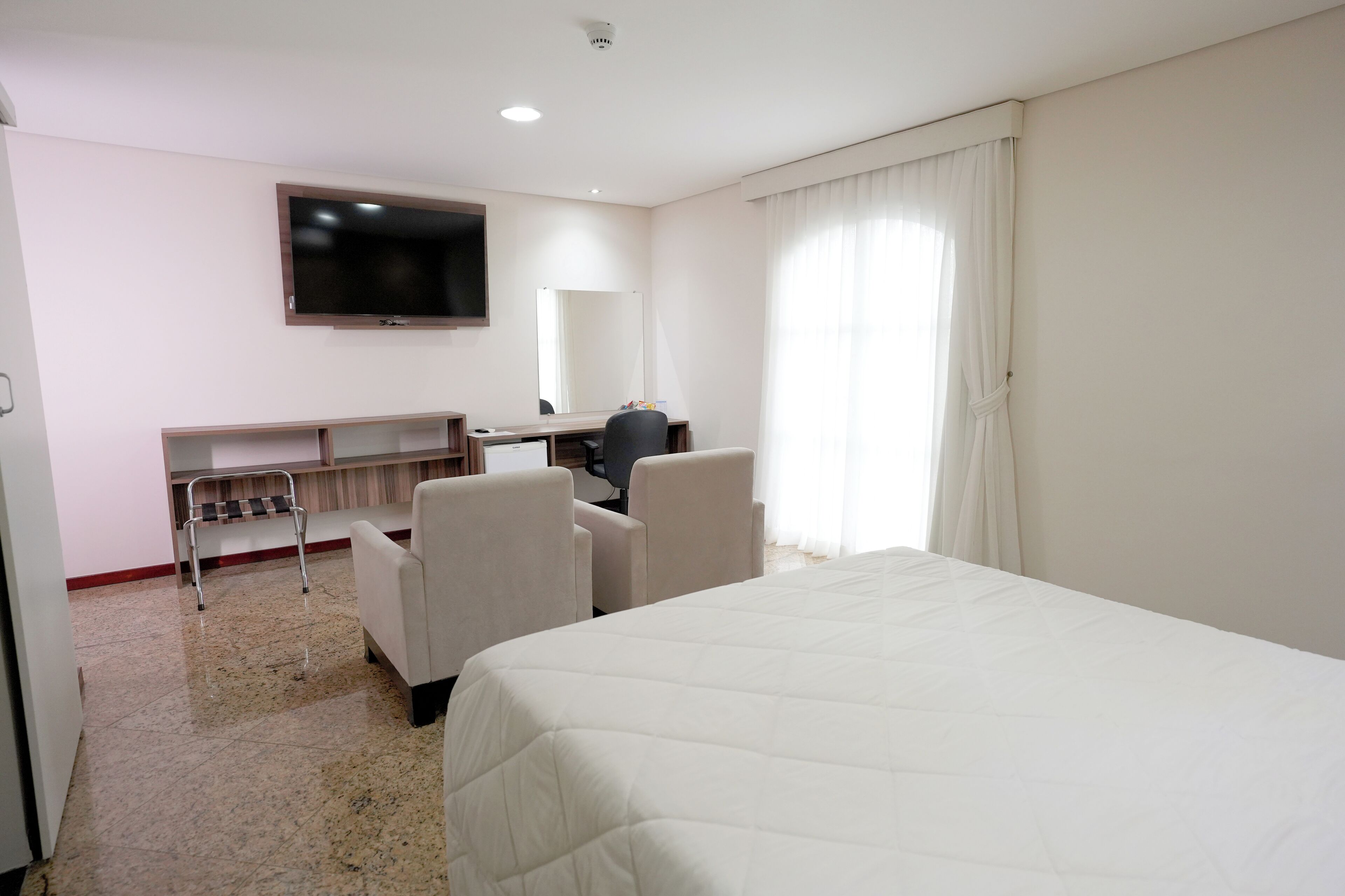 Quarto de hotel com cama, escrivaninha, cadeira, televisão e janela com cortinas.