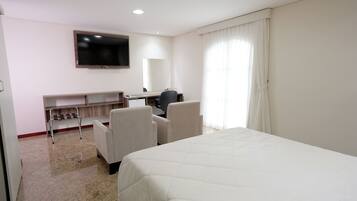 Quarto de hotel com cama, escrivaninha, cadeira, televisão e janela com cortinas.