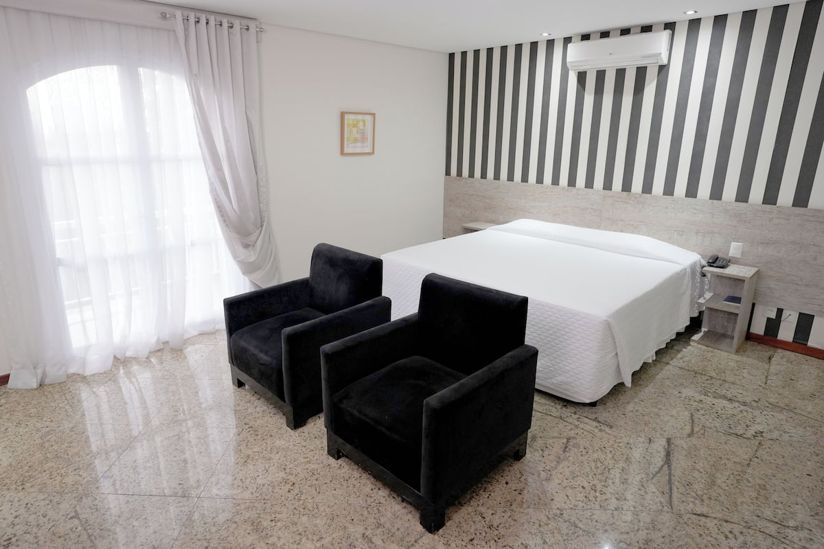 Une chambre d’hôtel avec un grand lit, deux fauteuils noirs, une table de chevet et un climatiseur fixé au mur.