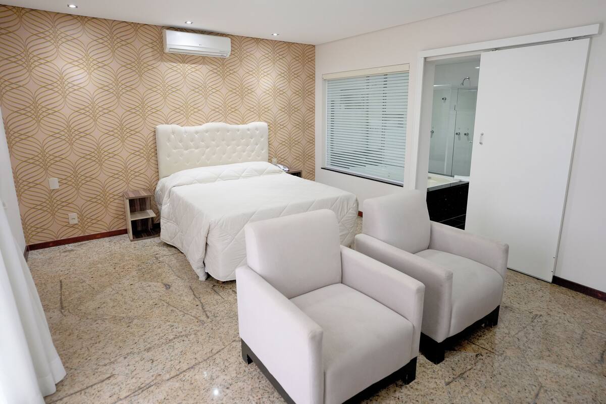 Une chambre à coucher comprenant un lit, deux fauteuils, une table de chevet, un miroir et une porte coulissante donnant sur une salle de bain.