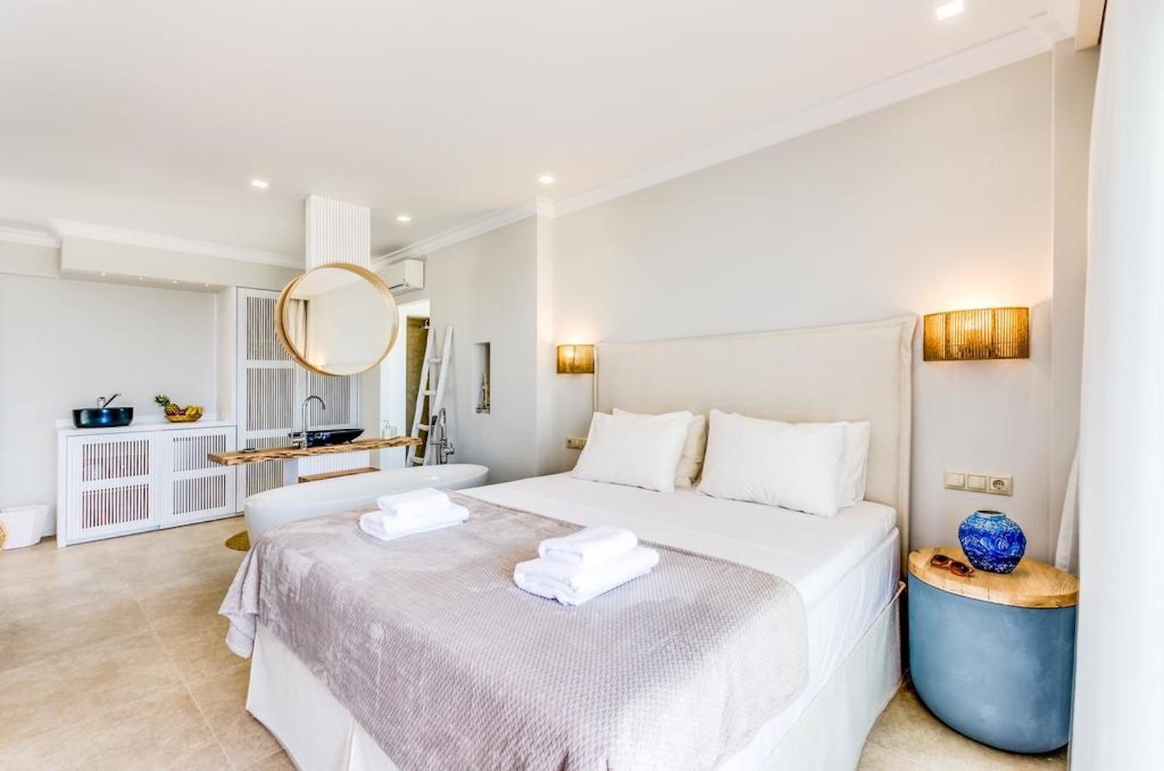 A modern bedroom with a large bed, a bedside table, a round mirror, and a blue vase.