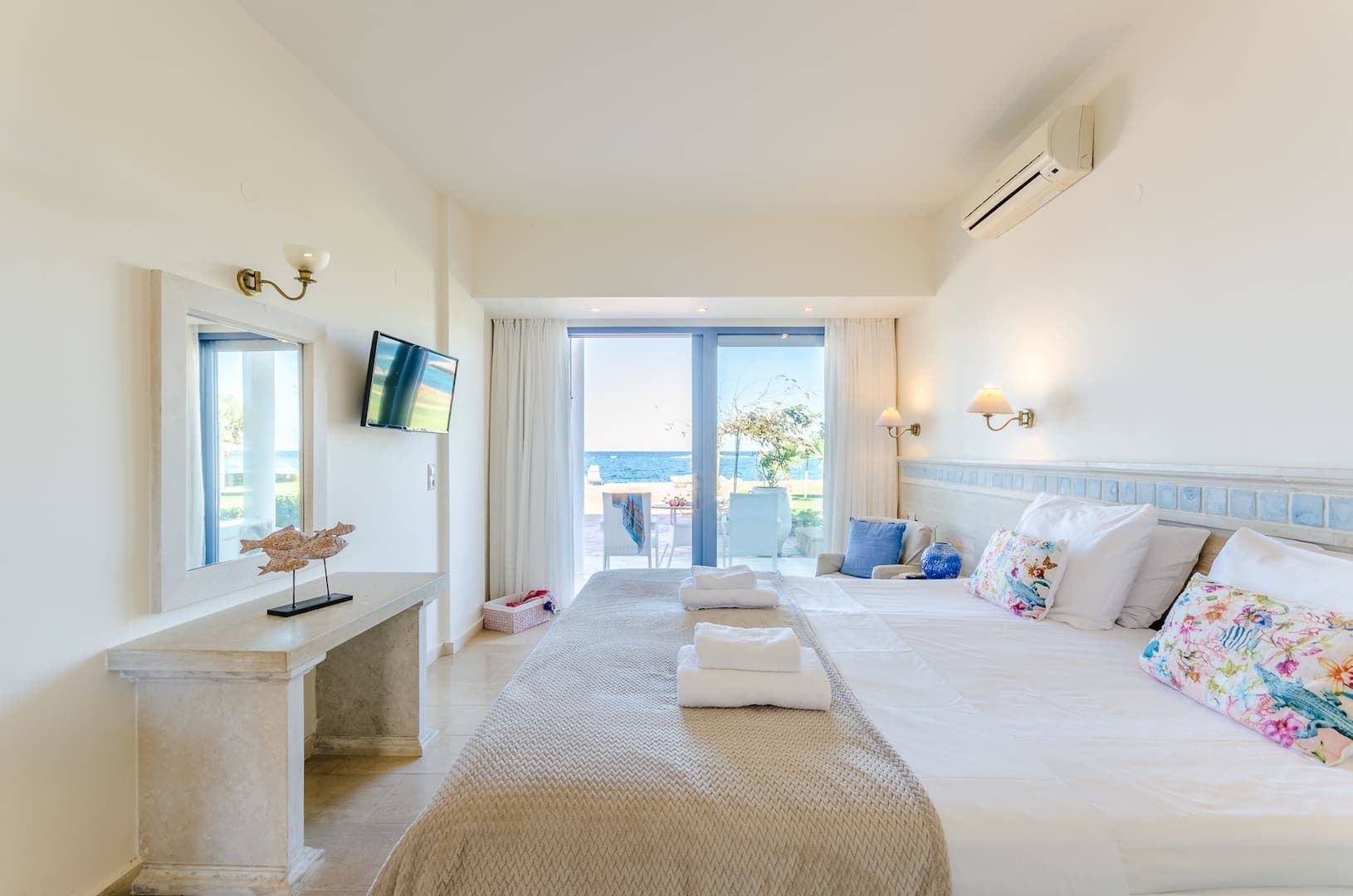 A hotel room with a bed, a desk, a TV, and a view of the sea.