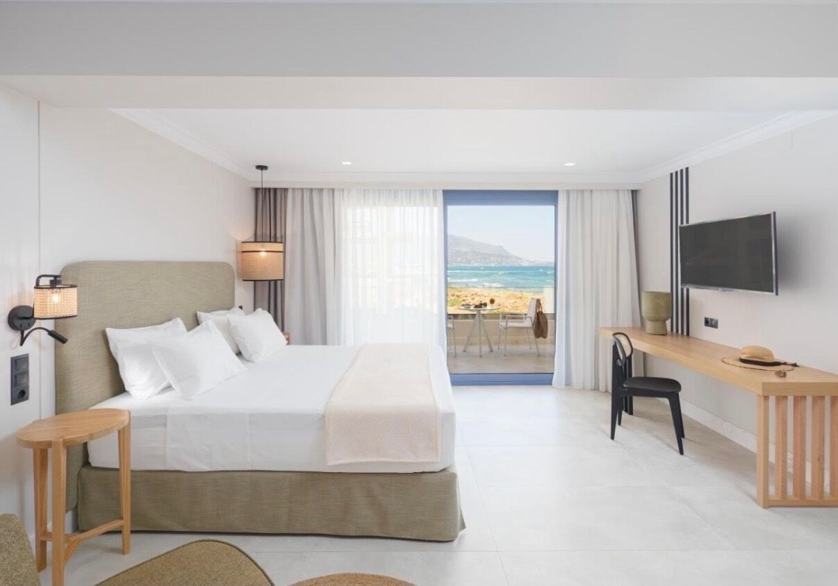 A hotel room with a bed, a desk, a chair, a TV, and a view of the beach.