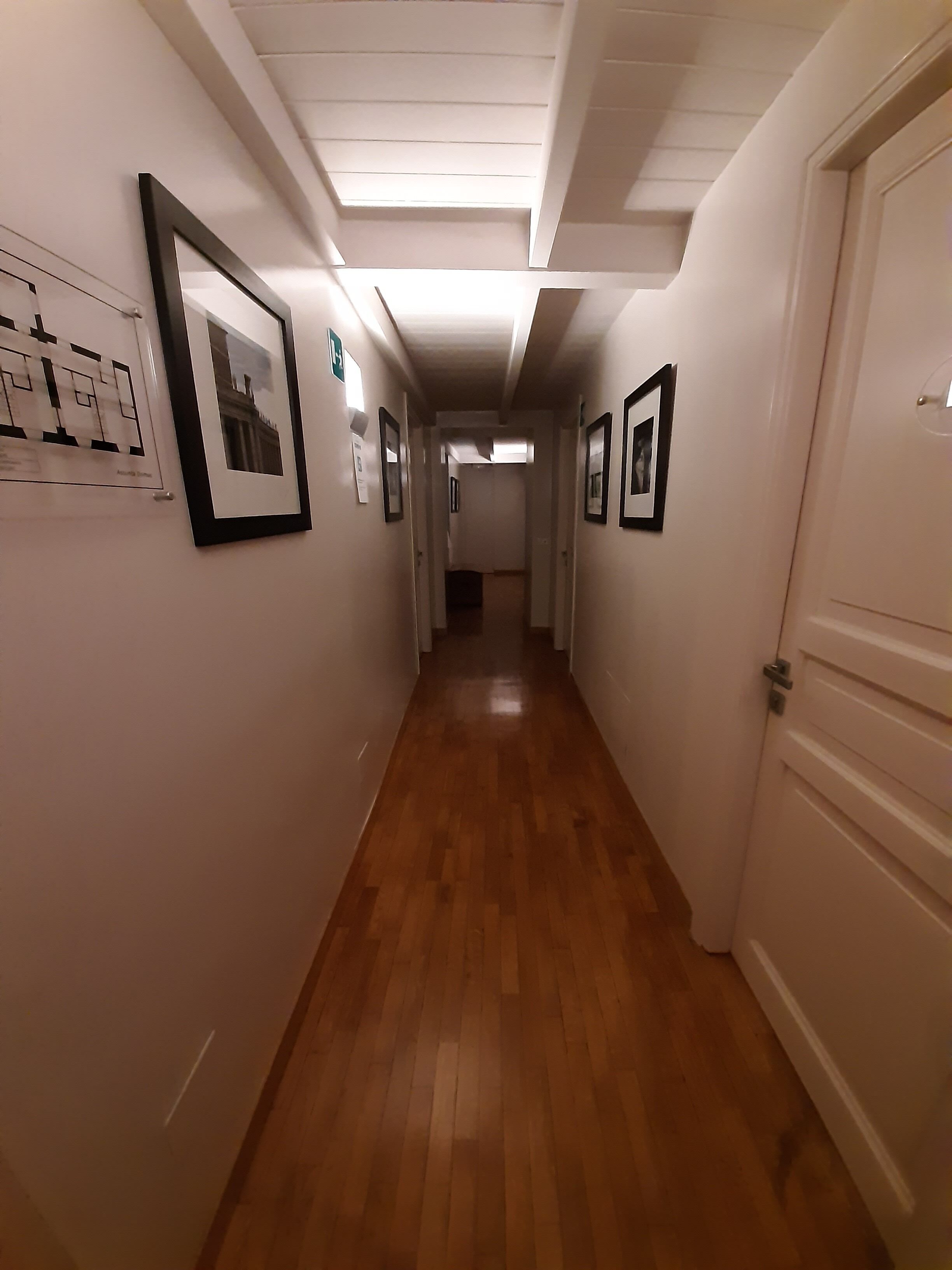 A hallway with framed pictures on the walls, wooden flooring, and a white door at the end.