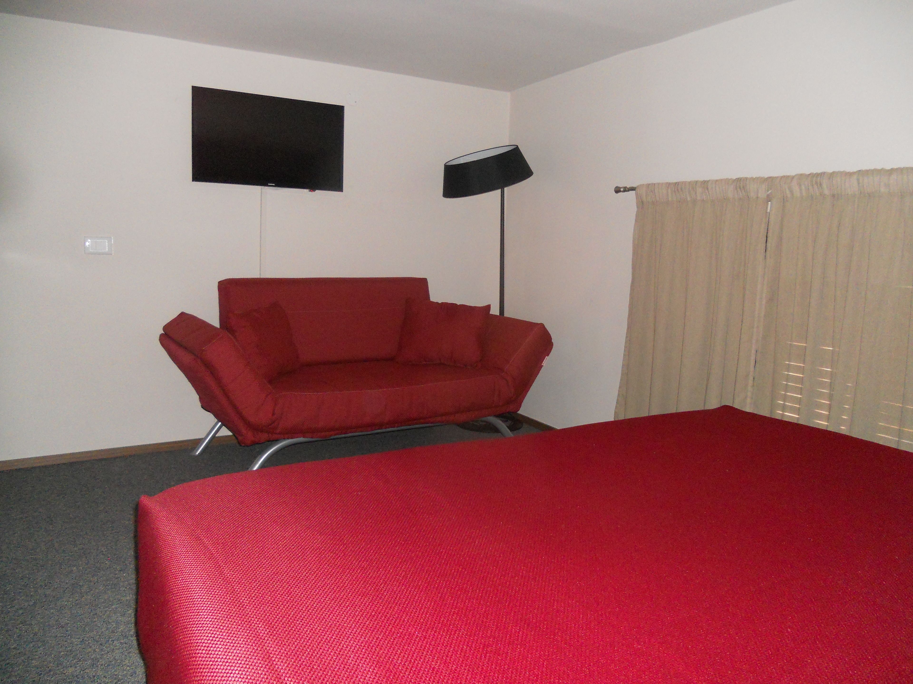 A room with a red sofa, a bed with a red cover, a flat-screen TV on the wall, and a floor lamp.