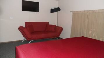 A room with a red sofa, a bed with a red cover, a flat-screen TV on the wall, and a floor lamp.