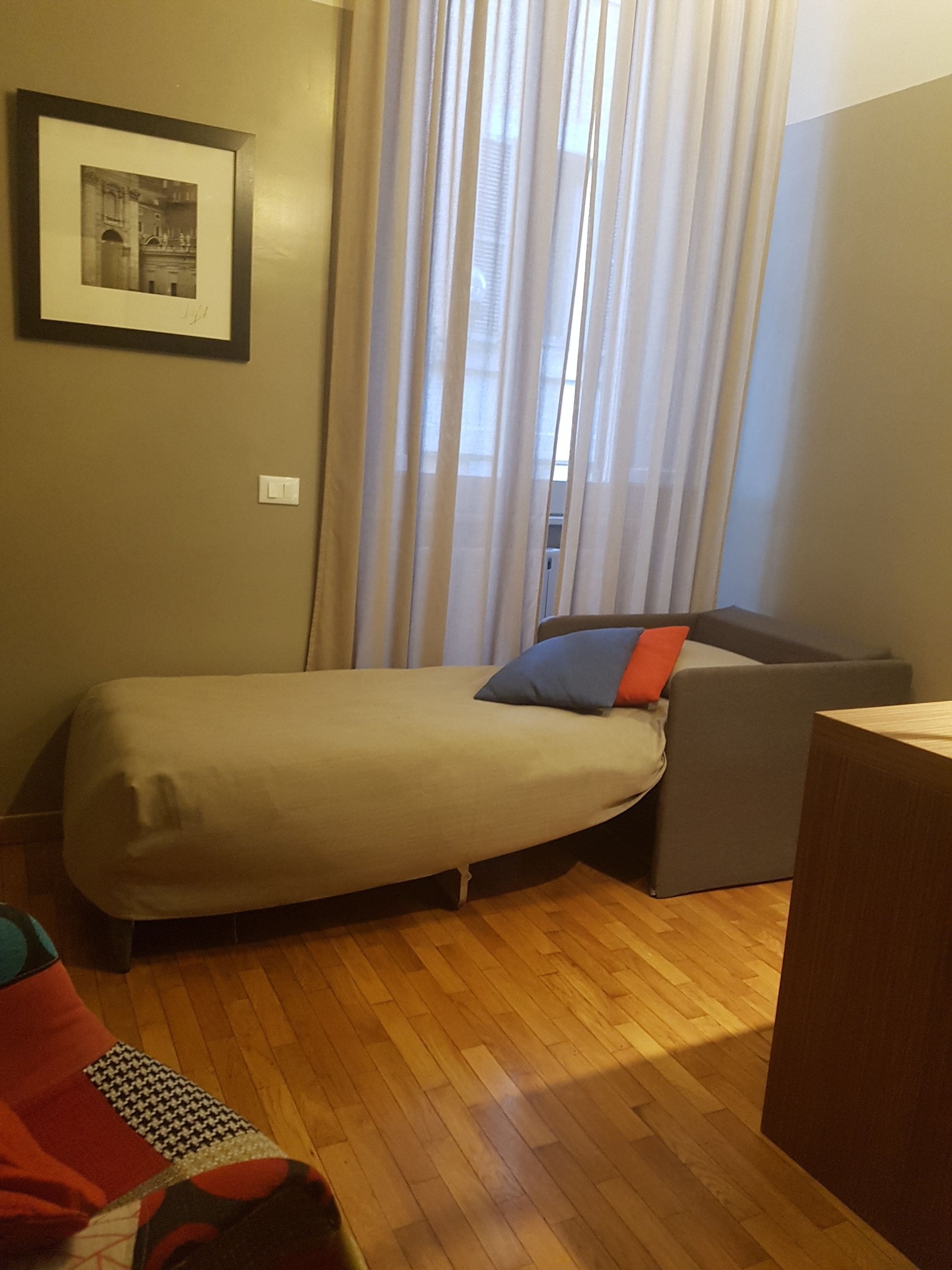 Une petite chambre avec un lit simple, un plancher en bois, un lit doté d’un matelas gris et d’un oreiller bleu et rouge, un canapé gris, un bureau en bois et une fenêtre avec des rideaux légers.