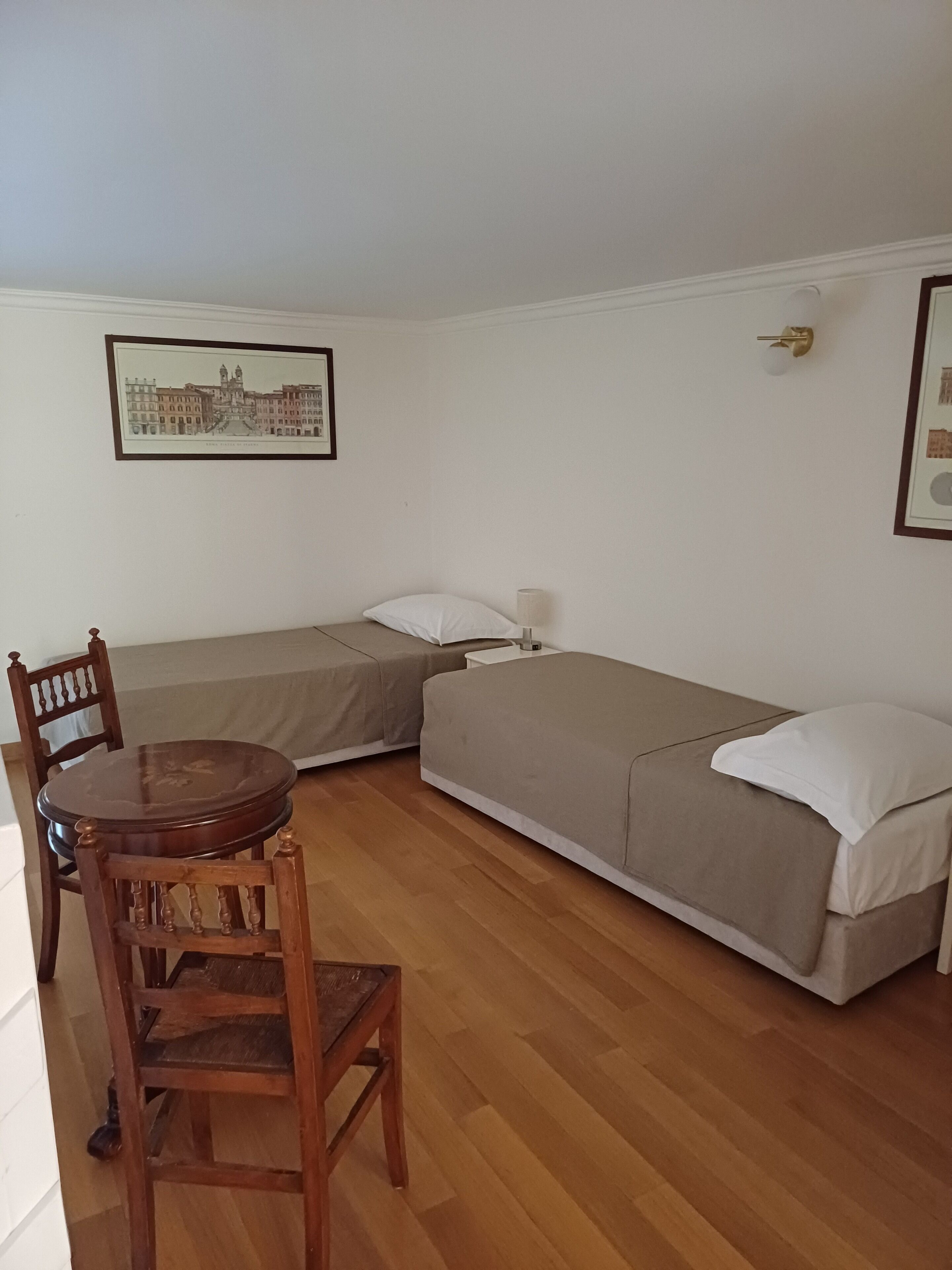 Een kleine, eenpersoons accommodatie met een houten vloer, een houten eettafel met vier stoelen, een enkel bed met een grijze matras, en een nachtkastje met een lamp.