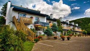 A motel with a parking lot, a staircase leading to the entrance, and a large tree in the foreground. - Motel Le JoliBourg (Saint-Sauveur)