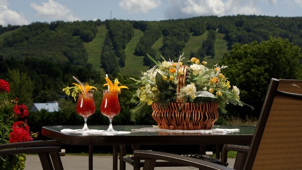 A table with two drinks and a floral arrangement, set against a backdrop of green hills. - Motel Le JoliBourg (Saint-Sauveur)
