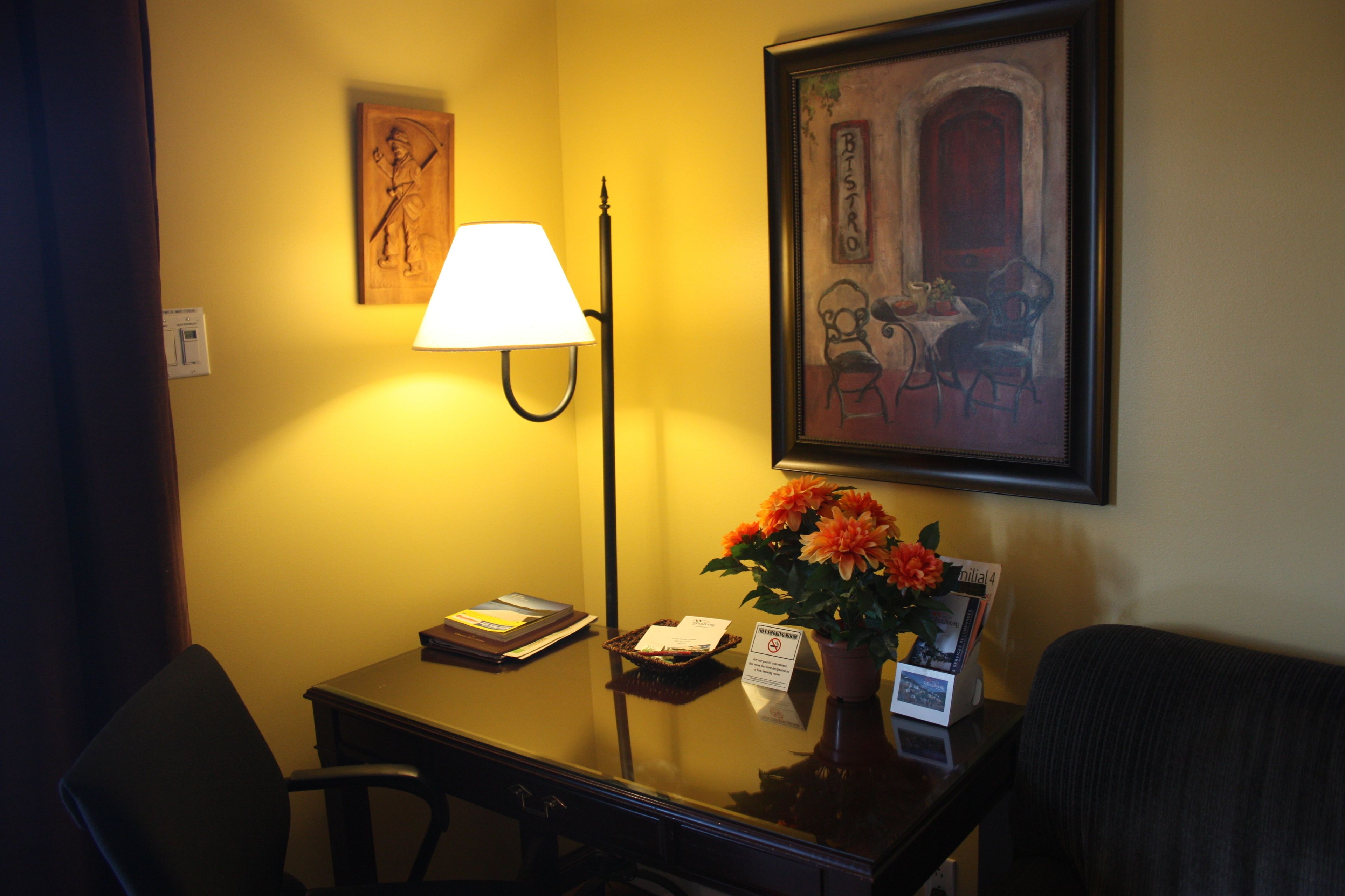 Una habitación de hotel con un escritorio, una lámpara, un cuadro enmarcado y un jarrón con flores.